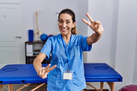 Photo for Young hispanic woman wearing physiotherapist uniform standing at clinic smiling looking to the camera showing fingers doing victory sign. number two. - Royalty Free Image