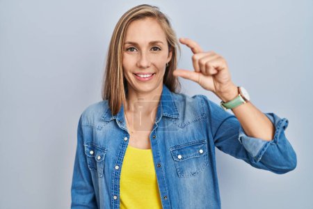Téléchargez les photos : Young blonde woman standing over blue background smiling and confident gesturing with hand doing small size sign with fingers looking and the camera. measure concept. - en image libre de droit