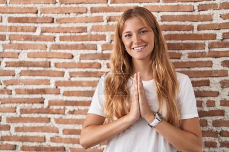 Photo for Young caucasian woman standing over bricks wall praying with hands together asking for forgiveness smiling confident. - Royalty Free Image