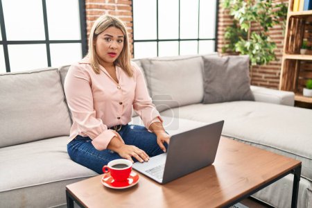 Photo for Young hispanic woman using laptop sitting on the sofa at home afraid and shocked with surprise expression, fear and excited face. - Royalty Free Image