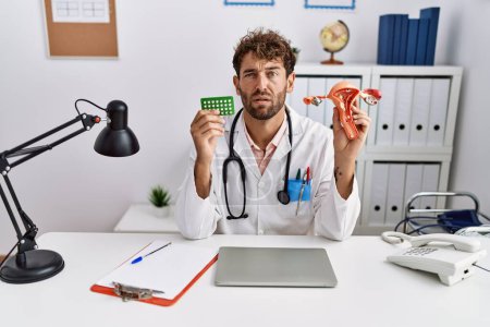 Photo for Young hispanic doctor man holding female genital organ and birth control pills clueless and confused expression. doubt concept. - Royalty Free Image