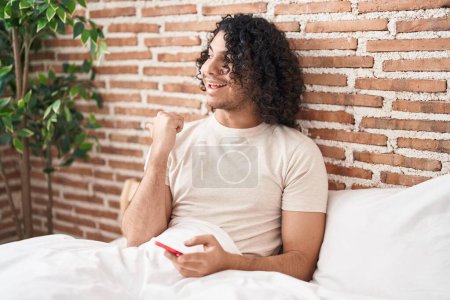 Téléchargez les photos : Homme hispanique avec les cheveux bouclés en utilisant smartphone assis sur le lit pointant le pouce vers le côté souriant heureux avec la bouche ouverte - en image libre de droit
