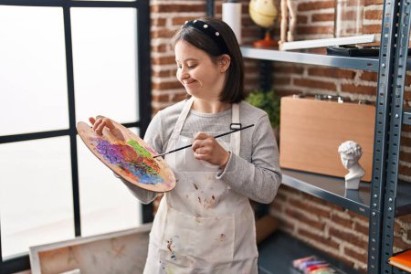 Photo for Young woman with down syndrome smiling confident holding paintbrush and palette at art studio - Royalty Free Image