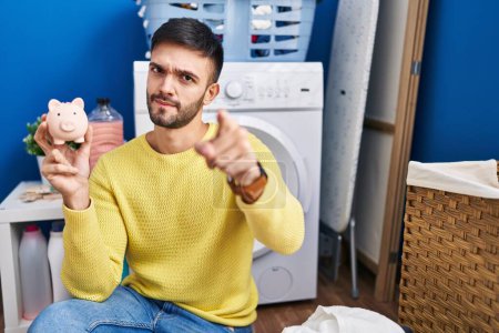 Foto de Hispanic man doing laundry holding piggy bank pointing with finger to the camera and to you, confident gesture looking serious - Imagen libre de derechos