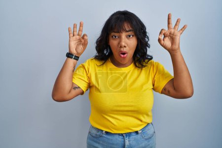 Photo for Hispanic woman standing over blue background looking surprised and shocked doing ok approval symbol with fingers. crazy expression - Royalty Free Image