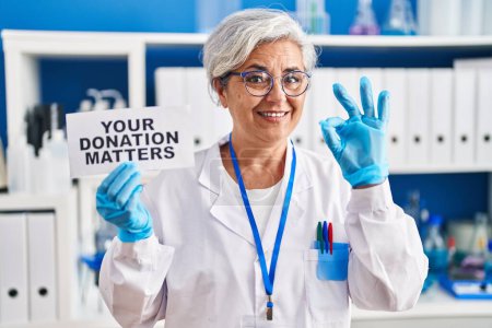 Téléchargez les photos : Femme d'âge moyen avec les cheveux gris travaillant au laboratoire scientifique tenant votre don questions bannière faire ok signe avec les doigts, sourire geste amical excellent symbole - en image libre de droit