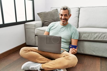 Téléchargez les photos : Homme aux cheveux gris d'âge moyen utilisant un ordinateur portable et carte de crédit assis sur le sol à la maison - en image libre de droit