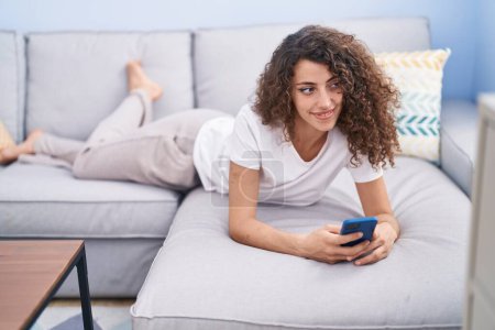 Foto de Mujer hispana hermosa joven usando teléfono inteligente tumbado en el sofá en casa - Imagen libre de derechos