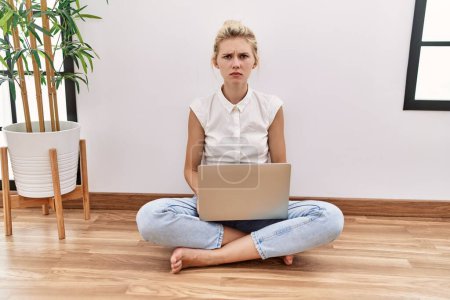 Foto de Mujer rubia joven usando ordenador portátil sentado en el suelo en la sala de estar escéptico y nervioso, frunciendo el ceño molesto debido a un problema. persona negativa. - Imagen libre de derechos