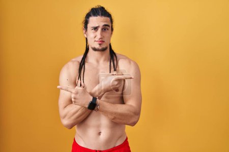 Téléchargez les photos : Homme hispanique avec les cheveux longs debout torse nu sur fond jaune pointant vers les deux côtés avec les doigts, direction différente en désaccord - en image libre de droit