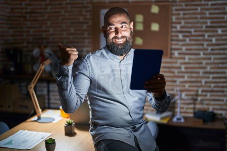 Téléchargez les photos : Jeune homme hispanique avec barbe et tatouages travaillant au bureau la nuit souriant avec le visage heureux regardant et pointant vers le côté avec le pouce vers le haut. - en image libre de droit