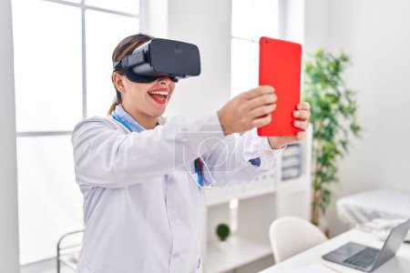 Photo for Young beautiful hispanic woman doctor using virtual reality glasses and touchpad at clinic - Royalty Free Image