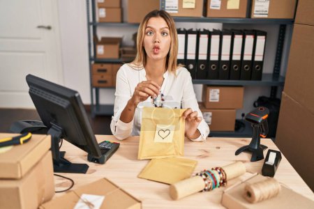 Foto de Young blonde woman working at small business ecommerce making fish face with mouth and squinting eyes, crazy and comical. - Imagen libre de derechos