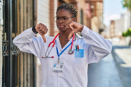 Téléchargez les photos : Afro-américaine femme médecin souriant confiant faire geste négatif avec les pouces vers le bas à la rue - en image libre de droit