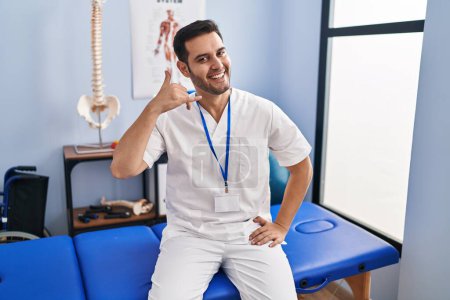 Photo for Young hispanic man with beard working at pain recovery clinic smiling doing phone gesture with hand and fingers like talking on the telephone. communicating concepts. - Royalty Free Image