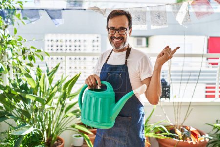Téléchargez les photos : Homme d'âge moyen portant tablier de jardinier tenant arrosoir à la maison terrasse souriant heureux pointant avec la main et le doigt sur le côté - en image libre de droit