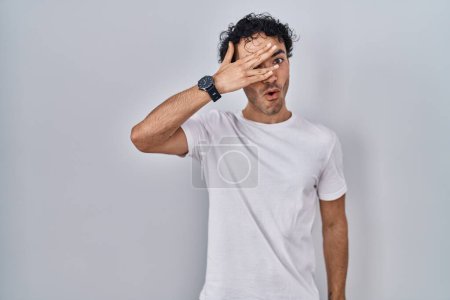 Foto de Hombre hispano de pie sobre un fondo aislado asomándose en conmoción cubriendo cara y ojos con la mano, mirando a través de los dedos con expresión avergonzada. - Imagen libre de derechos