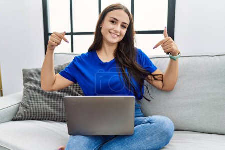 Foto de Mujer morena joven utilizando el ordenador portátil en casa buscando confiado con sonrisa en la cara, señalándose con los dedos orgullosos y felices. - Imagen libre de derechos
