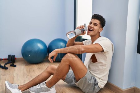 Photo for Young hispanic man drinking water sitting on floor at sport center - Royalty Free Image