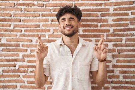 Téléchargez les photos : Arab man with beard standing over bricks wall background gesturing finger crossed smiling with hope and eyes closed. luck and superstitious concept. - en image libre de droit