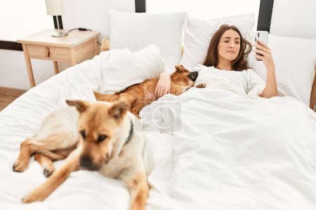 Foto de Joven mujer hispana abrazando perro y usando smartphone acostado en la cama en el dormitorio - Imagen libre de derechos
