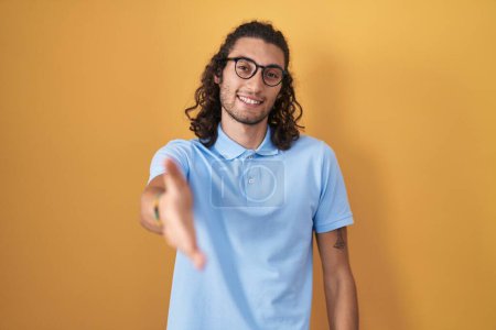 Foto de Joven hombre hispano de pie sobre fondo amarillo sonriente amistoso ofreciendo apretón de manos como saludo y bienvenida. negocios exitosos. - Imagen libre de derechos