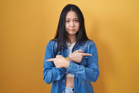 Téléchargez les photos : Jeune femme asiatique debout sur fond jaune pointant vers les deux côtés avec les doigts, direction différente en désaccord - en image libre de droit