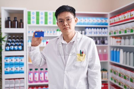 Téléchargez les photos : Jeune homme asiatique travaillant à la pharmacie pharmacie tenant carte de crédit pensée attitude et expression sobre regardant confiance en soi - en image libre de droit