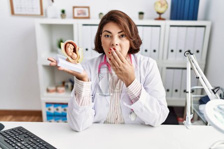 Photo for Middle age hispanic doctor woman holding anatomical model of female uterus with fetus covering mouth with hand, shocked and afraid for mistake. surprised expression - Royalty Free Image
