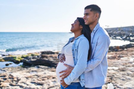 Foto de Young latin couple expecting baby hugging each other standing at seaside - Imagen libre de derechos