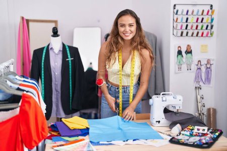 Foto de Young beautiful hispanic woman tailor smiling confident cutting cloth at tailor shop - Imagen libre de derechos