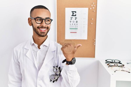 Téléchargez les photos : African american optician man standing by eyesight test smiling with happy face looking and pointing to the side with thumb up. - en image libre de droit