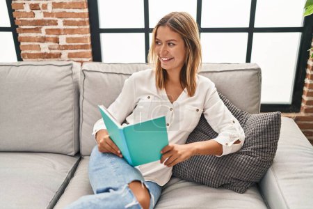 Photo for Young hispanic woman reading book sitting on sofa at home - Royalty Free Image