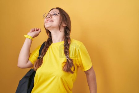 Foto de Joven mujer caucásica usando mochila de estudiante sobre fondo amarillo bailando feliz y alegre, sonriendo moviéndose casual y confiado escuchando música - Imagen libre de derechos