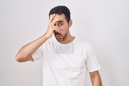 Foto de Handsome hispanic man standing over white background peeking in shock covering face and eyes with hand, looking through fingers with embarrassed expression. - Imagen libre de derechos