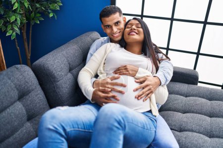 Foto de Joven pareja latina esperando un bebé tocando el vientre tumbado en el sofá en casa - Imagen libre de derechos