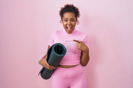 Foto de Young hispanic woman with curly hair holding yoga mat over pink background surprised pointing with finger to the side, open mouth amazed expression. - Imagen libre de derechos