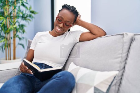 Foto de African american woman reading book sitting on sofa at home - Imagen libre de derechos