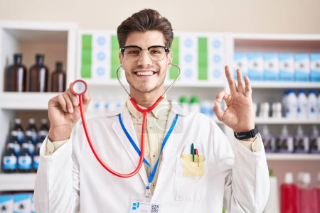 Foto de Young hispanic man working at pharmacy drugstore using stethoscope doing ok sign with fingers, smiling friendly gesturing excellent symbol - Imagen libre de derechos