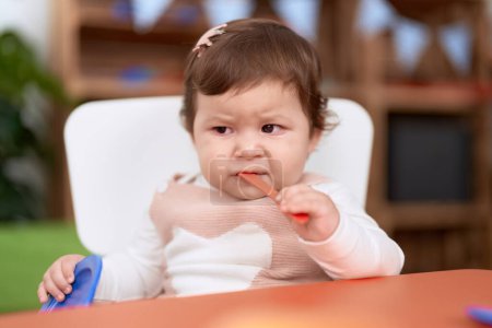 Foto de Adorable niño sentado en la mesa chupando cuchara de plástico sosteniendo el plato en el jardín de infantes - Imagen libre de derechos