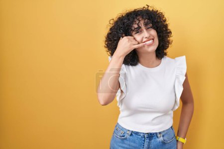 Photo for Young middle east woman standing over yellow background smiling doing phone gesture with hand and fingers like talking on the telephone. communicating concepts. - Royalty Free Image