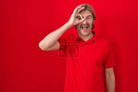 Foto de Caucasian man with mustache standing over red background doing ok gesture with hand smiling, eye looking through fingers with happy face. - Imagen libre de derechos