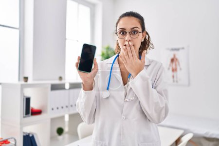 Téléchargez les photos : Jeune médecin hispanique femme tenant smartphone montrant écran couvrant la bouche avec la main, choqué et peur de l'erreur. expression surprise - en image libre de droit