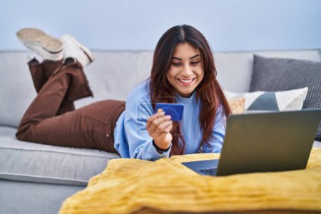 Photo for Young hispanic woman using laptop and credit card lying on sofa at home - Royalty Free Image