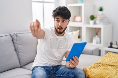 Photo for Hispanic man with beard using touchpad sitting on the sofa pointing with finger surprised ahead, open mouth amazed expression, something on the front - Royalty Free Image