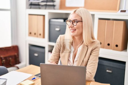 Téléchargez les photos : Jeune femme blonde travailleuse d'affaires utilisant un ordinateur portable et des écouteurs au bureau - en image libre de droit