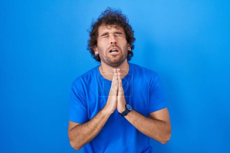 Photo for Hispanic young man standing over blue background begging and praying with hands together with hope expression on face very emotional and worried. begging. - Royalty Free Image