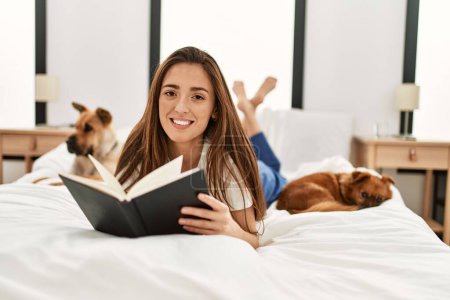 Photo for Young hispanic woman reading book lying on bed with dogs at bedroom - Royalty Free Image