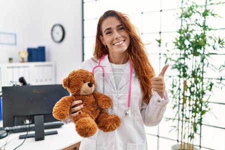 Téléchargez les photos : Jeune femme pédriaticienne rousse tenant ours en peluche souriant heureux et positif, pouce levé faire excellent et signe d'approbation - en image libre de droit