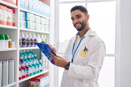 Téléchargez les photos : Jeune homme arabe pharmacien souriant écriture confiante sur presse-papiers à la pharmacie - en image libre de droit